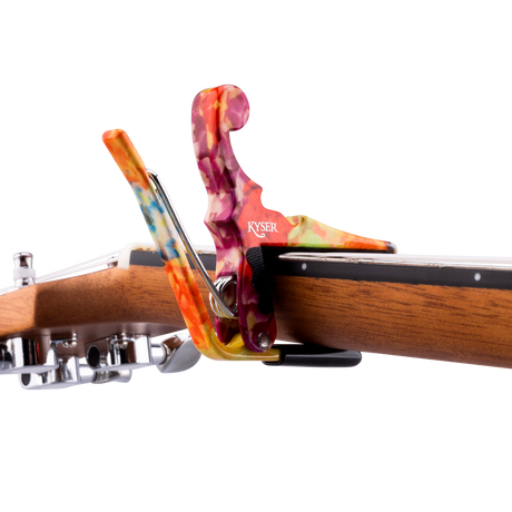 The image shows a Kyser guitar capo with a vibrant, swirled design in orange, purple, pink, and other colors, attached to the neck of an acoustic guitar. The capo features a spring-loaded mechanism for quick, one-handed use. Its colorful, tie-dye style finish adds a decorative touch to this essential accessory that clamps the fretboard to raise the pitch and enable easy key changes while maintaining chord fingerings.