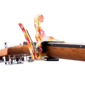 The image shows a Kyser spring-loaded clamp capo attached to the neck of an acoustic guitar. The capo features a vibrant, marbled pattern with swirls of pink, orange, yellow, and white, creating a colorful, tie-dye-like appearance. Part of the guitar’s wooden neck, fretboard, headstock, and tuning pegs are visible, highlighting both the capo’s functionality and eye-catching design.