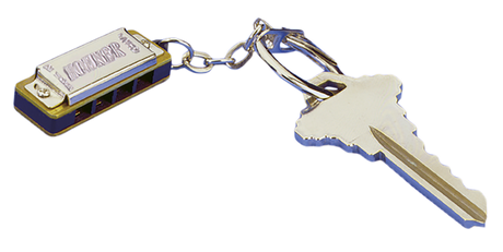 The image shows a miniature harmonica keychain attached to a key ring with a house key. The harmonica is small and functional, with a metallic silver finish and blue accents. It has a classic rectangular shape with holes on the front and the word "HARMONICA" embossed on the top. This novelty item serves as both a playable instrument and a keychain, popular as a compact musical accessory or gift.