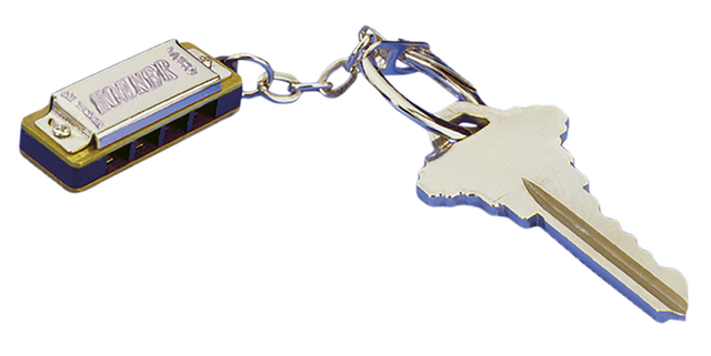 The image shows a miniature harmonica keychain attached to a key ring with a house key. The harmonica is small and functional, with a metallic silver finish and blue accents. It has a classic rectangular shape with holes on the front and the word "HARMONICA" embossed on the top. This novelty item serves as both a playable instrument and a keychain, popular as a compact musical accessory or gift.