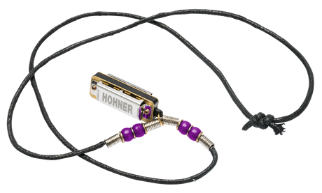 The image shows a Hohner harmonica transformed into a wearable necklace. The small, chrome harmonica with gold trim features the Hohner logo and is suspended on a black braided cord. Purple beads flank the instrument, connected by metal coils, creating a handcrafted, artistic look. This unique accessory allows musicians to keep their harmonica handy while making a stylish statement.