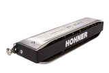 The image shows a Hohner harmonica with a black body featuring "HOHNER" in white letters, a chrome top plate with engraved text, and multiple front holes. This compact diatonic wind instrument is made by a renowned German brand since 1857, commonly used in blues, folk, country, and rock. Sound is produced as air flows through the holes, vibrating metal reeds inside.