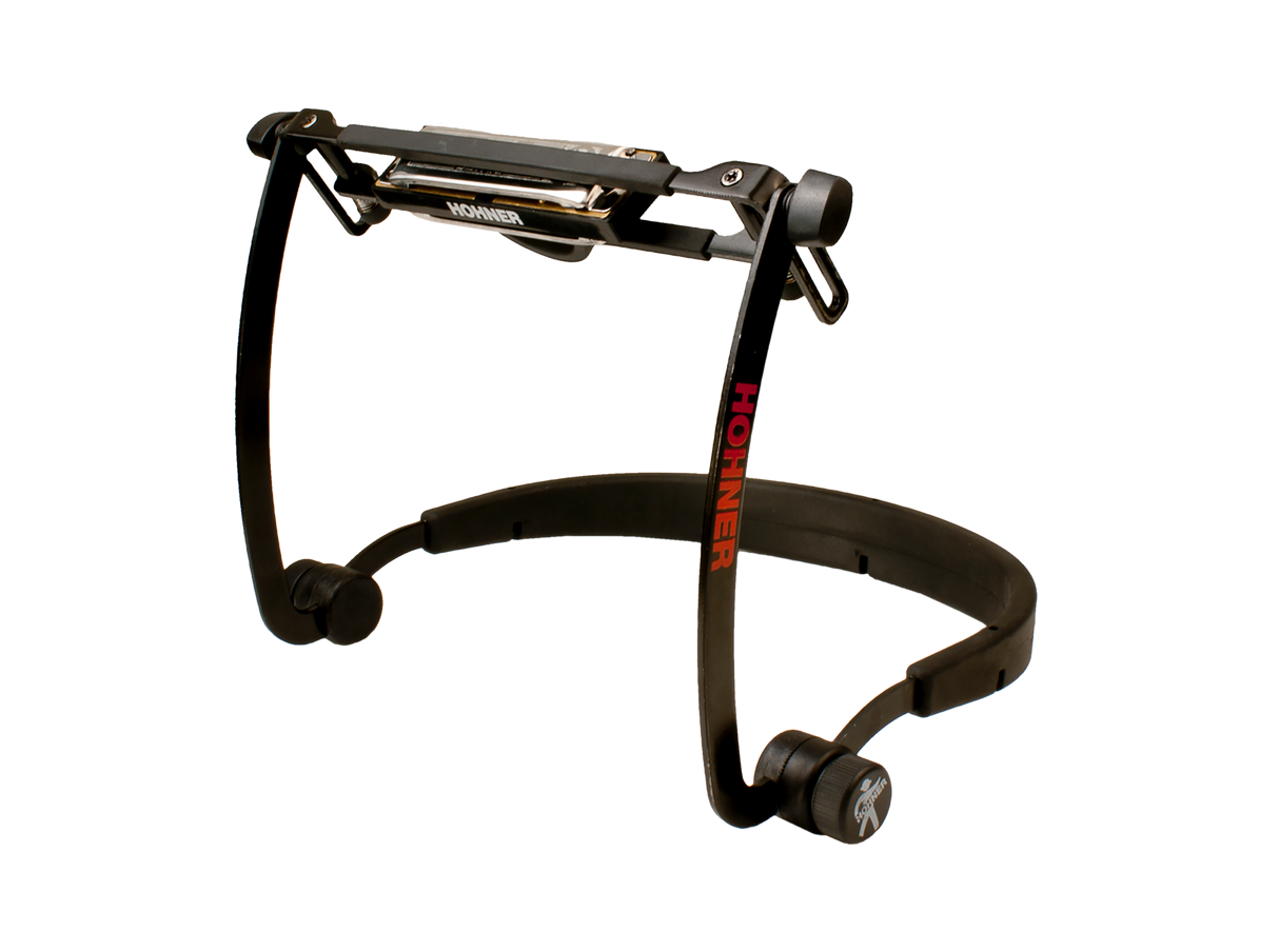 The image shows a Hohner harmonica holder with a black metal frame and adjustable padded neck strap for hands-free play. The holder secures the harmonica, displays the "HOHNER" brand, and allows tilting for customized positioning. Commonly used by folk and blues musicians, it enables playing harmonica while playing other instruments. The sturdy, professional design suits practice and live performance.