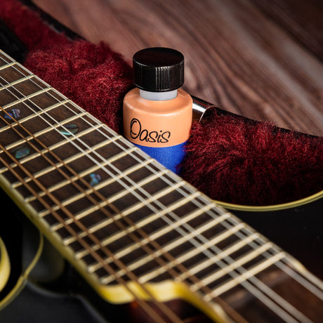 The image shows an acoustic guitar with a dark wood body on a wooden surface, alongside an Oasis guitar humidifier bottle with an orange cap and a burgundy cleaning cloth. This setup highlights essential guitar maintenance tools, focusing on humidity control to protect acoustic guitars from damage due to dry conditions.