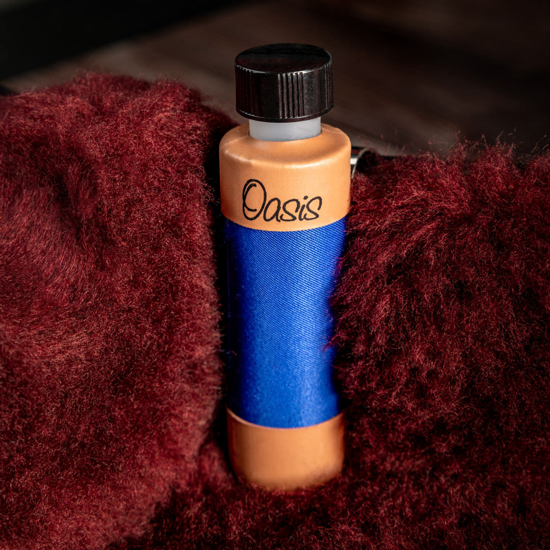 The image shows an "Oasis" humidifier, a guitar accessory for maintaining humidity in wooden string instruments. It features a black screw-on cap, an orange/gold top and bottom with the "Oasis" logo, and a blue fabric middle section that holds moisture. The humidifier rests in red/burgundy fabric, likely inside a guitar case, helping prevent wood damage by controlling humidity levels to preserve the instrument’s condition.