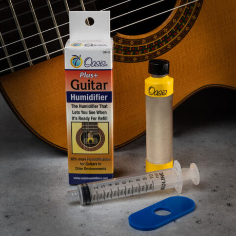 The image shows the Oasis OH-6 Plus+ Guitar Humidifier system placed in front of an acoustic guitar. The kit includes a cylindrical humidifier tube, a small bottle of yellow humidifying solution, a syringe for filling, a blue plastic clip, and the original packaging. The humidifier fits in the guitar’s sound hole to maintain proper humidity, protecting the wood from cracking and warping.