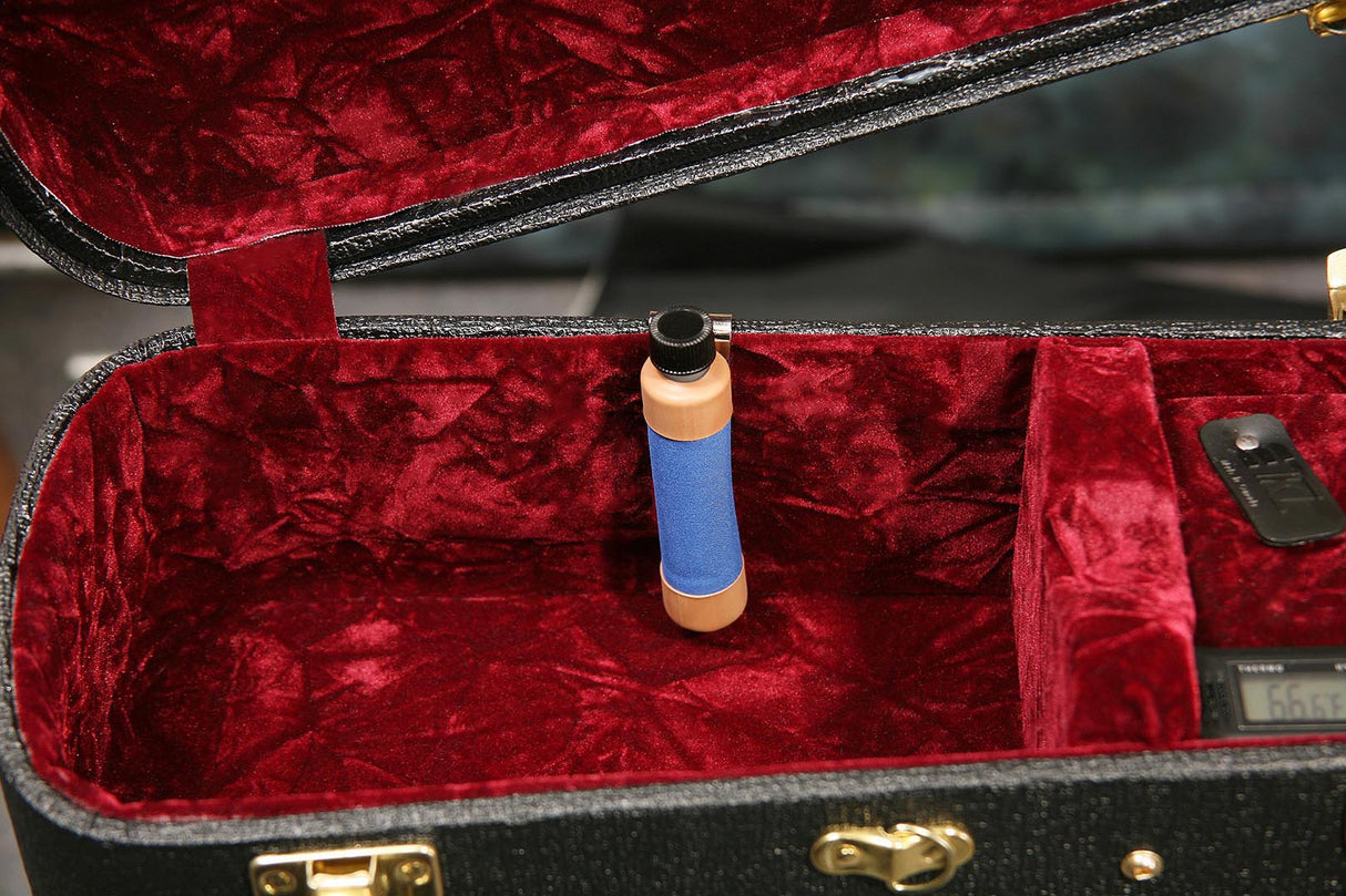 The image shows a professional microphone inside a black hard case with deep red velvet lining. The microphone features a blue fabric windscreen, gold/brass metal body sections, a black grille, and a threaded mount at the bottom. The case includes foam padding and a small electronic accessory. This setup suggests high-quality audio equipment used for recording or live performance.