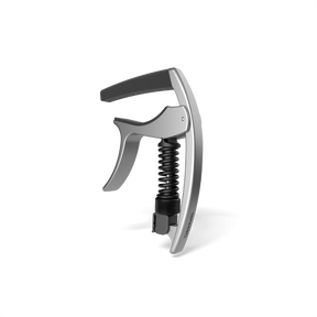 The image shows a guitar capo with a sleek silver metallic body and a black ergonomic handle. It features a visible black coiled spring in its spring-loaded, trigger-style release system for quick one-handed use. Designed to clamp the fretboard evenly, it raises string pitch without harming the guitar. Suitable for acoustic and electric guitars, this precision capo helps musicians easily change keys while maintaining chord fingerings.