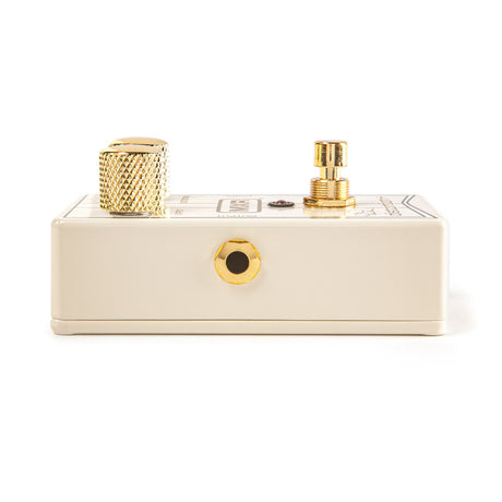 The image shows a compact cream-colored guitar effects pedal with gold/brass hardware. It features two prominent knurled gold knobs on top—one standard control knob and one that may be a stomp switch. A gold output jack is visible on the front. The pedal has some unreadable labeling on top. Its high-quality gold hardware suggests it is a boutique or premium model used to modify guitar sounds with effects like overdrive or boost.