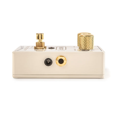 The image shows a cream-colored guitar effects pedal with gold hardware. The top panel features a gold threaded input jack on the left, a large gold knurled knob on the right, and a small switch or control markings in the center. The side panel has two jacks, one black and one gold. The pedal is compact and sturdy, designed to shape guitar tones for both studio and live use.