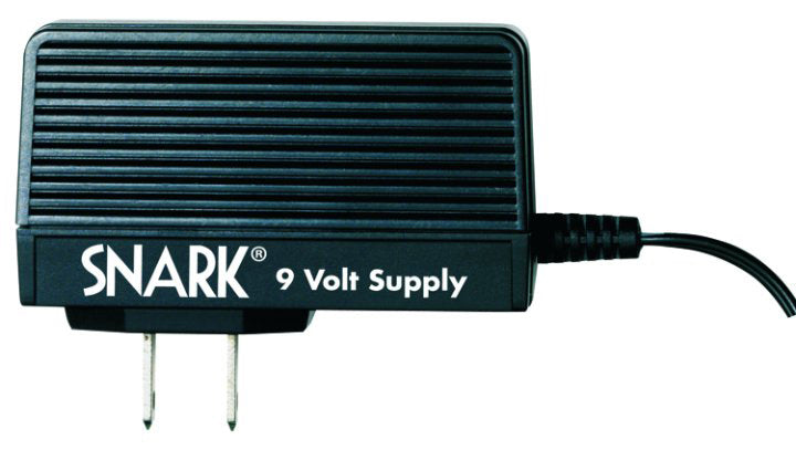 The image shows a black SNARK 9 Volt power supply adapter for musical instruments. It features "SNARK" and "9 Volt Supply" branding, wall plug prongs, and a power cable to connect electronic music gear. This adapter powers effects pedals, tuners, and other 9-volt devices, providing reliable power and eliminating the need for batteries.
