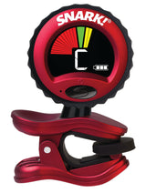 The image shows a Snark clip-on guitar tuner with a red and black design and the "SNARK!" logo. It has a circular LCD display with a multicolored arc indicating tuning status (flat, in tune, sharp) and shows the note "C." A clip at the bottom attaches to the instrument's headstock, and a power button is visible on the front. This popular tuner picks up vibrations for accurate tuning of guitars and other stringed instruments.