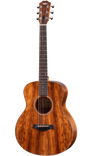 The image shows a beautiful Taylor steel-string acoustic guitar with a medium-sized Grand Auditorium body. It features a rich koa or mahogany wood grain with amber tones, chrome tuning machines, a dark wood bridge with a compensated saddle, a traditional round soundhole with a decorative rosette, and a dark ebony or rosewood fretboard with dot markers. This high-quality instrument is suited for recording, performing, or serious playing.