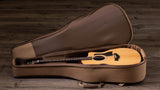 The image shows a dreadnought-style acoustic guitar with a light wood top and darker wood sides resting in an open brown hard or semi-hard case with a plush interior. The case has custom-fitted compartments for the guitar’s body and neck. The setup is photographed against a rustic wooden background, highlighting a warm, traditional aesthetic typical of acoustic guitar storage and transport.