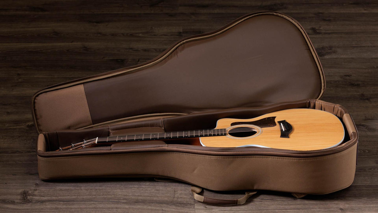 The image shows an acoustic steel-string guitar with a natural wood finish resting inside an open brown/tan hard or semi-hard case with plush lining. The guitar has a light-colored top, darker back and sides, a sound hole, bridge, and dark fretboard. The case’s fitted interior securely holds the guitar. The scene is set on a wooden floor with a rustic background, creating a warm, musical atmosphere.