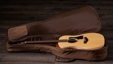 The image shows an acoustic guitar with a natural wood finish resting in a padded, tan soft case with a plush interior and zippered closure. The guitar features a dreadnought body, round sound hole, and dark pickguard. The case is placed on a rustic dark wooden surface, creating a warm, vintage look typical of product photography for guitars and accessories.