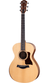 The image shows a Taylor acoustic guitar with a mid-size body, natural spruce top, and darker wood back and sides. It features a round soundhole with a decorative rosette, dark tortoiseshell pickguard, dark wood bridge with white pins, and a dark fretboard with dot markers. The headstock has Taylor’s signature shape with chrome tuners. This mid-to-upper range guitar is known for quality craftsmanship and balanced tone.