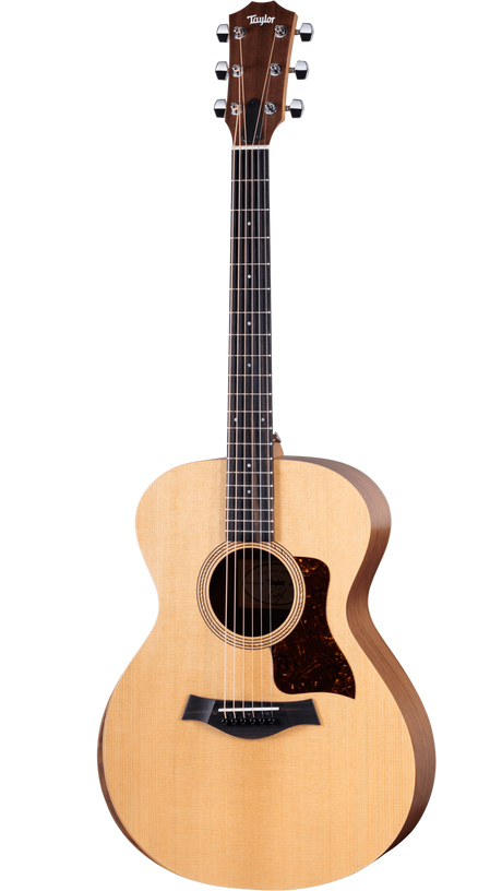 The image shows a Taylor acoustic guitar with a mid-size body, natural spruce top, and darker wood back and sides. It features a round soundhole with a decorative rosette, dark tortoiseshell pickguard, dark wood bridge with white pins, and a dark fretboard with dot markers. The headstock has Taylor’s signature shape with chrome tuners. This mid-to-upper range guitar is known for quality craftsmanship and balanced tone.