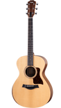 The image shows a Taylor acoustic guitar with a mid-size body, natural spruce top, and darker wood back and sides. It features a round soundhole with a decorative rosette, dark tortoiseshell pickguard, dark wood bridge with white pins, and a dark fretboard with dot markers. The headstock has Taylor’s signature shape with chrome tuners. This mid-to-upper range guitar is known for quality craftsmanship and balanced tone.