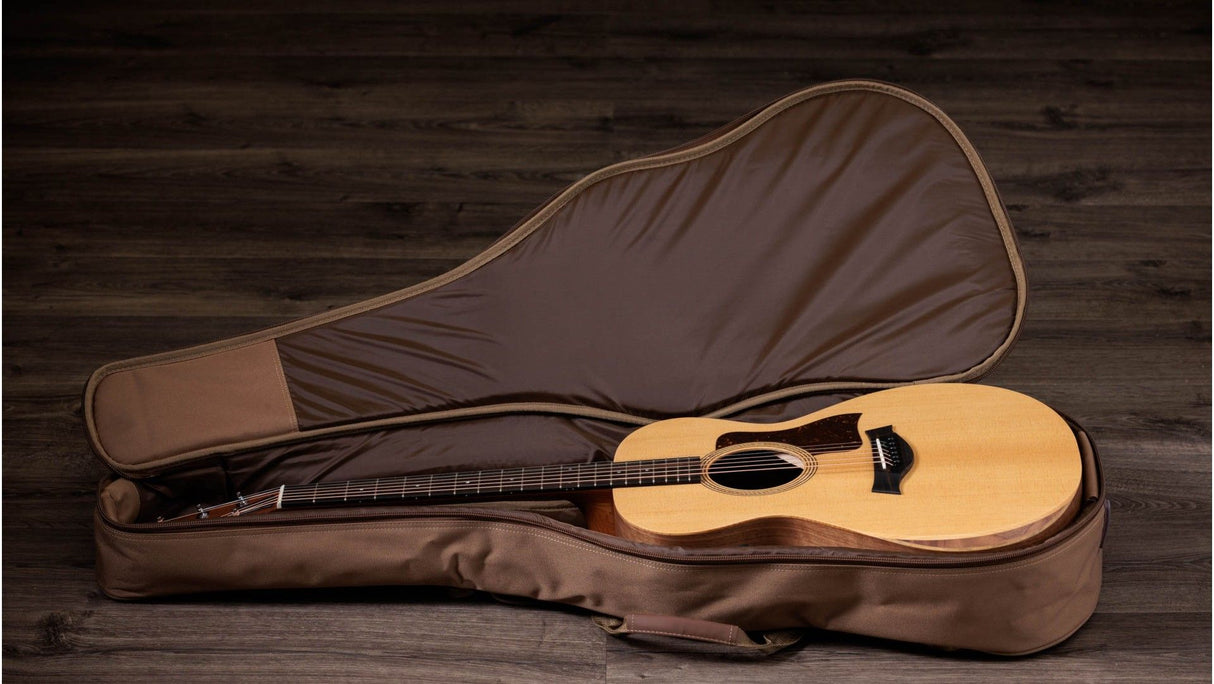 The image shows an acoustic guitar with a natural light wood finish and dark fretboard resting in an open, brown soft-shell case with plush interior lining. The guitar, likely a steel-string dreadnought, sits on a dark wooden surface, contrasting with the warm tones of the instrument and case, illustrating typical portable storage and transport gear used by musicians.