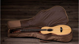 The image shows an acoustic steel-string guitar with a natural wood finish resting inside an open brown padded gig bag. The guitar has a light-colored soundboard and darker back and sides, featuring a dreadnought or similar body shape. The gig bag has a plush interior lining and a small accessory pocket. The background is rustic wood, creating a warm, traditional atmosphere typical of acoustic guitar displays.