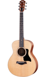 The image shows a Taylor steel-string acoustic guitar with a natural spruce or cedar top and darker mahogany or sapele back and sides. It features a mid-sized Grand Auditorium body, chrome tuning machines, a dark wood bridge with a compensated saddle, a round soundhole with a rosette, and a black pickguard. The natural finish highlights the wood grain, reflecting Taylor’s quality craftsmanship and versatile playability.