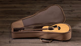 The image shows an acoustic guitar with a natural wood finish resting in an open tan soft-shell case. The guitar has a traditional body shape and a dark fretboard with inlays. The case features a plush, dark interior lining and lies open on a weathered wooden surface, providing padded protection for the instrument during transport.