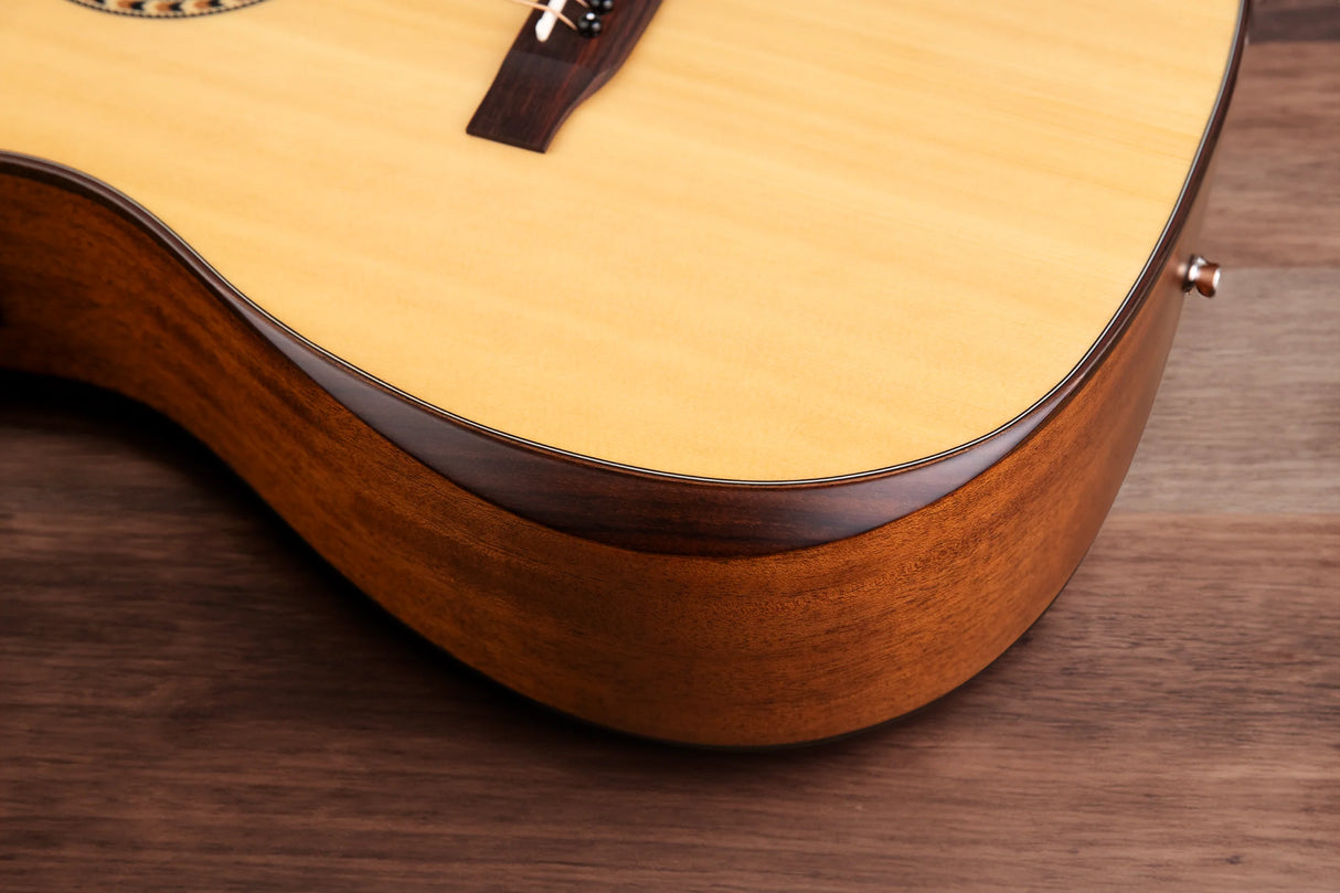 The image shows a close-up of the lower bout of an acoustic guitar, highlighting a light-colored spruce or cedar soundboard with natural finish, dark wood grain back and sides, dark binding along the edges, part of the wooden bridge, and a partial view of the rosette around the sound hole. The guitar rests on a wooden surface, showcasing fine craftsmanship and contrasting tones typical of quality acoustic guitars.