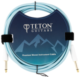 The image shows a Teton Guitars Premium Woven Instrument Cable with a light blue/turquoise woven fabric exterior for durability and style. It has gold-plated 1/4" connectors on both ends for optimal signal transfer. The packaging features the Teton Guitars logo and mountain design. The cable is coiled and secured with velcro straps, highlighting its quality build ideal for electric guitars and basses.