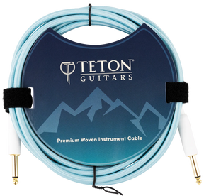 The image shows a Teton Guitars Premium Woven Instrument Cable with a light blue/turquoise woven fabric exterior for durability and style. It has gold-plated 1/4" connectors on both ends for optimal signal transfer. The packaging features the Teton Guitars logo and mountain design. The cable is coiled and secured with velcro straps, highlighting its quality build ideal for electric guitars and basses.