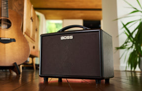 The image shows a compact BOSS guitar amplifier with a dark gray finish and prominent branding on the front grille. It has a sturdy handle and sits on a wooden floor. In the background, a partially visible acoustic guitar complements the setup. The scene appears to be a cozy home music room or studio with warm lighting and plants. BOSS is known for quality guitar amps and pedals, and this model is suitable for practice or small performances.