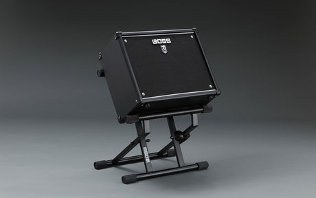 The image shows a compact black BOSS guitar amplifier mounted on an adjustable BOSS-branded stand. The amp features the distinctive BOSS logo on its front grille and is tilted for optimal sound direction. This portable setup is ideal for guitarists needing elevated amplification for better sound projection and easy control access, commonly used by both amateur and professional musicians.