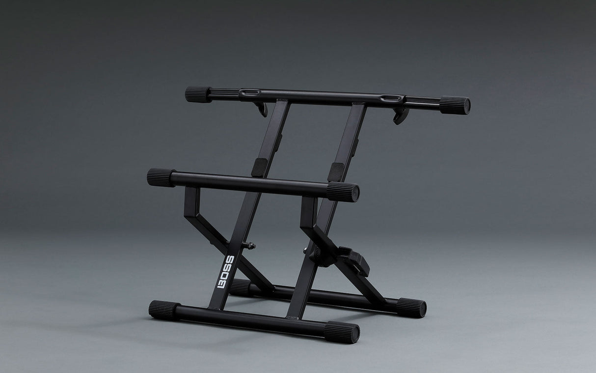 The image shows a black Boss adjustable keyboard stand with a sturdy metal X-frame base. It features a dual-tier design with two horizontal support rails, adjustable height and angle settings, rubber end caps to prevent slipping, and a foldable structure for easy transport and storage. This stand is commonly used by keyboardists and electronic musicians to position instruments ergonomically for practice, recording, or live performance.
