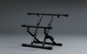 The image shows a black Boss adjustable keyboard stand with a sturdy metal X-frame base. It features a dual-tier design with two horizontal support rails, adjustable height and angle settings, rubber end caps to prevent slipping, and a foldable structure for easy transport and storage. This stand is commonly used by keyboardists and electronic musicians to position instruments ergonomically for practice, recording, or live performance.