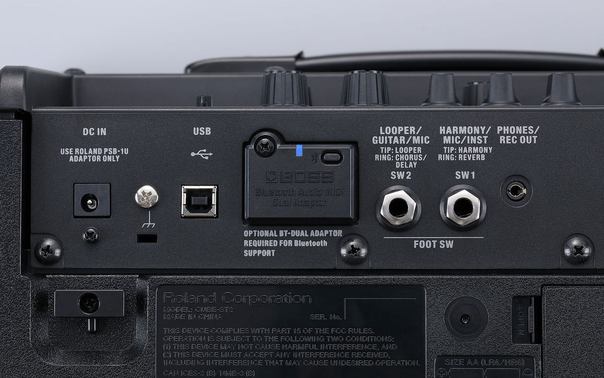 The image shows the back panel of a Roland CUBE-ST2 guitar/bass amplifier featuring a DC IN jack, USB port, various input/output jacks (LOOPER/GUITAR/MIC, HARMONY/MIC/INST, PHONES/REC OUT), SW1 and SW2 function switches, FOOT SW connection, a slot for BOSS Bluetooth Audio MIDI Dual Adaptor, battery compartment for AA batteries, and compliance labels. It highlights the amp's blend of analog and digital features, including built-in effects and wireless connectivity.