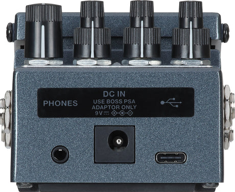 The image shows a BOSS guitar effects pedal with a blue/gray metal body, featuring eight control knobs in two rows, input/output jacks, a headphone output labeled "PHONES," a USB port, and a power input for a 9V BOSS PSA adapter. Designed as a stomp-box, it allows musicians to activate effects by foot and is used to modify and enhance the sound of electric guitars, basses, and other instruments.