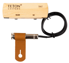 The image shows a TETON Guitars THM2 acoustic guitar pickup and preamp system. It includes a light wood preamp/control box with "VOLUME" and "MIC" controls, a black pickup cable, a chrome 1/4" output jack, and a brown leather mounting strap. Designed to amplify acoustic guitars, the hybrid system combines pickup and microphone functions, with a wooden preamp for an aesthetic fit inside the guitar.