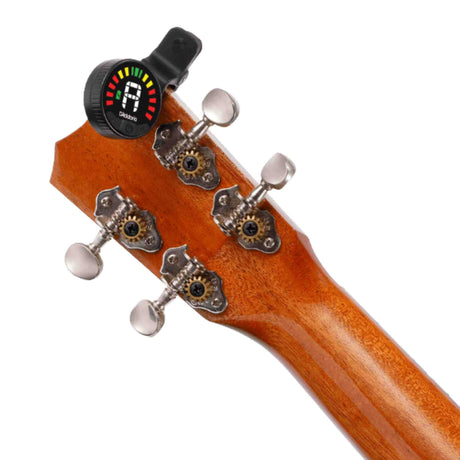 The image shows the headstock of a stringed instrument, likely a guitar or bass, with a clip-on electronic tuner attached. The wooden headstock has an amber finish and four chrome tuning pegs with white buttons. The black tuner displays a colorful LED semicircle and shows the note "A." This popular accessory reads vibrations to help musicians tune strings accurately, even in noisy environments.