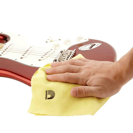 The image shows a hand using a yellow D'Addario polishing cloth to clean a reddish-burgundy electric guitar with a white pickguard. This microfiber cloth is a guitar maintenance accessory used to remove fingerprints and keep the instrument’s finish clean. D'Addario is a well-known brand that produces guitar strings, picks, and care products, helping musicians preserve their instruments' appearance and quality.