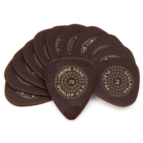 The image shows a collection of dark brown, tortoiseshell Dunlop guitar picks with gold dotted text patterns. The picks feature "PRIMETONE" and ".73," indicating the thickness gauge. These premium, vintage-inspired picks have elegant stippled lettering. Guitar picks are essential for plucking or strumming and come in various thicknesses and materials to suit different playing styles.