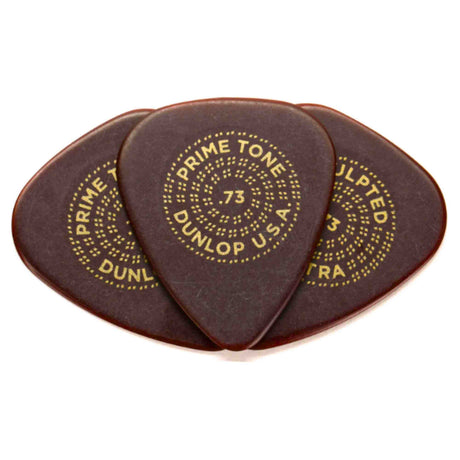 The image shows three dark brown or tortoiseshell Dunlop Primetone guitar picks with gold lettering. Each pick features "PRIME TONE," "DUNLOP U.S.A," and the number "73" (0.73mm thickness) arranged in a circular pattern. These premium teardrop-shaped picks are made from Primetone material, known for its smooth feel, warm tone, durability, and a textured grip area favored by acoustic and electric guitar players.