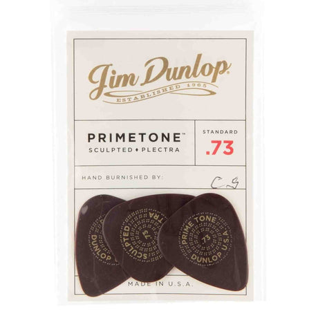 The image shows a package of three Jim Dunlop Primetone guitar picks, made in the USA. These sculpted plectra have a standard .73mm thickness (red "73" marking) and are hand burnished, featuring initials "C.G." Each dark-colored pick displays circular text including "PRIMETONE" and "DUNLOP." Known for premium tone and feel, these picks are favored by serious guitarists.