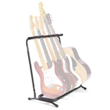 The image shows a black metal multi-guitar stand designed to hold several guitars, featuring a folding, portable A-frame or tripod base with padded contact points. Behind it are multiple guitars, including electric models like a pinkish Stratocaster-style guitar and a natural wood acoustic. This stand is commonly used by musicians, music stores, studios, teachers, and venues for display and easy transport.