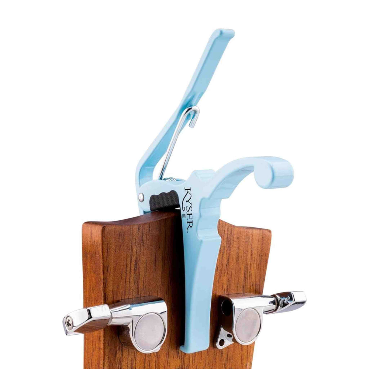 The image shows a light blue/turquoise Kyser Quick-Change guitar capo clamped on the neck of an acoustic guitar with visible natural wood and chrome tuning pegs. The capo, a popular accessory for guitarists, uses a spring-loaded mechanism for easy one-handed use, allowing players to raise the pitch of all strings by clamping across the fretboard. It’s widely used across many music genres.