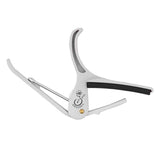 The image shows a Grover guitar capo with a sleek silver finish and ergonomic curved arms. It has a black padded bar that clamps all guitar strings and a spring-loaded mechanism with a gold-colored pivot. Designed for quick, one-handed use, this trigger-style capo allows guitarists to easily change keys by clamping across any fret while maintaining chord fingerings. The Grover logo is visible on its main body.
