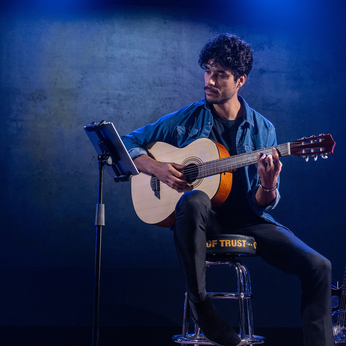 Man playing guitar on a stool while looking at a tablet with a dark blue background 