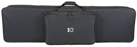 The image shows a Korg soft-shell keyboard gig bag in black fabric with a "K" logo on the front zippered pocket. It features a large main compartment for a keyboard, top carry handles, and a long rectangular shape. Designed for easy transport, it protects electronic keyboards from dust, moisture, and minor impacts while storing accessories like cables and pedals in the front pocket.