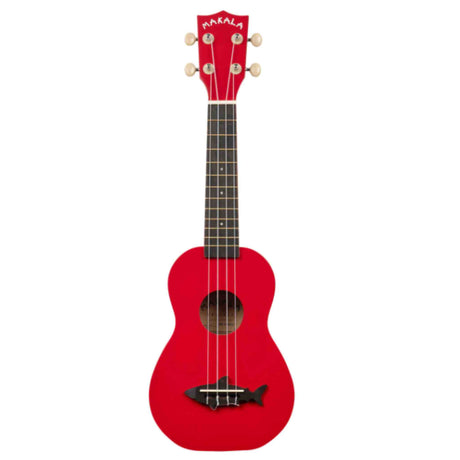 The image shows a bright red Makala soprano ukulele with a dark fretboard featuring dot markers, four strings, six tuning pegs with light-colored buttons, and a black bridge with a decorative shark design. The "MAKALA" logo is visible on the headstock. This small, traditional ukulele is popular among beginners for its affordability and vibrant, eye-catching appearance.