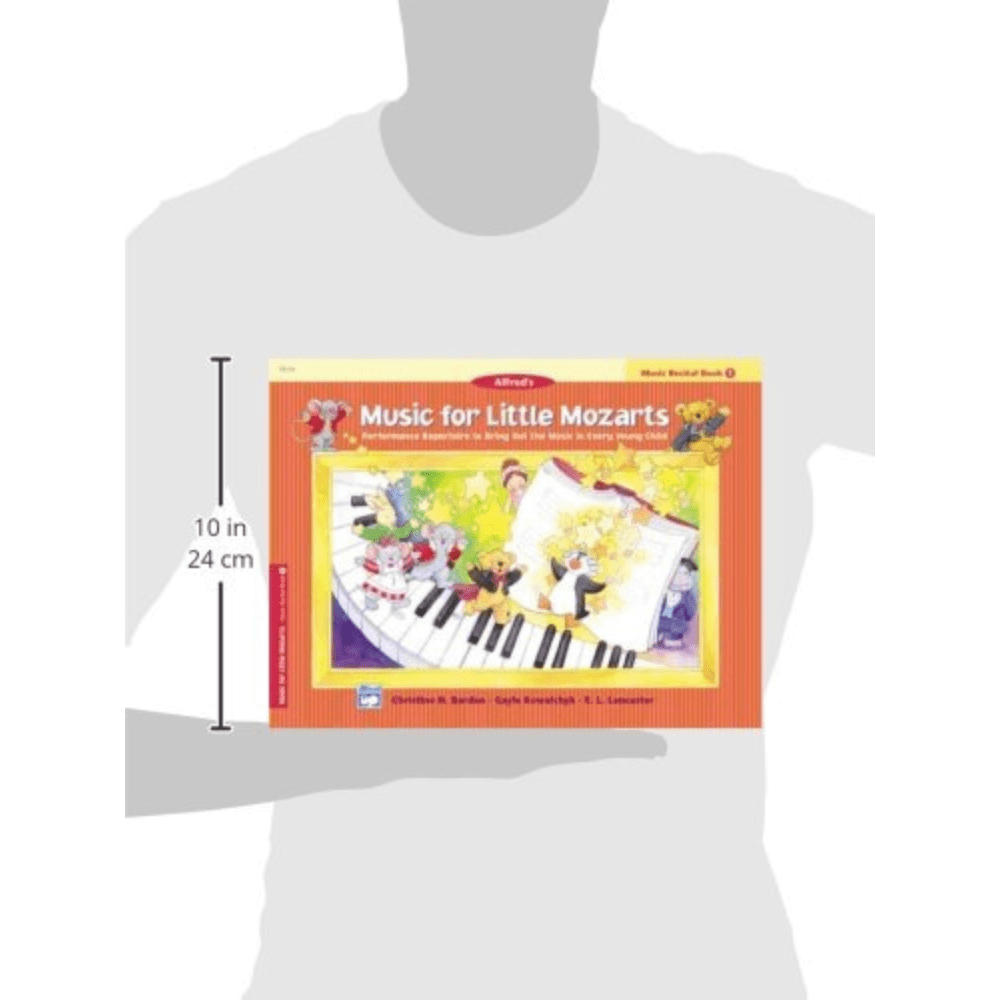 The image shows a person holding a colorful music education book titled "Music for Little Mozarts: Music Recital Book 1" by Alfred. The cover features cartoon children and musical characters around a piano keyboard, with bright orange and yellow tones. This beginner piano method book for young learners measures about 10 inches tall and includes simple pieces, basic theory, and engaging illustrations designed to make learning fun.