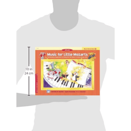 The image shows a person holding a colorful music education book titled "Music for Little Mozarts: Music Recital Book 1" by Alfred. The cover features cartoon children and musical characters around a piano keyboard, with bright orange and yellow tones. This beginner piano method book for young learners measures about 10 inches tall and includes simple pieces, basic theory, and engaging illustrations designed to make learning fun.